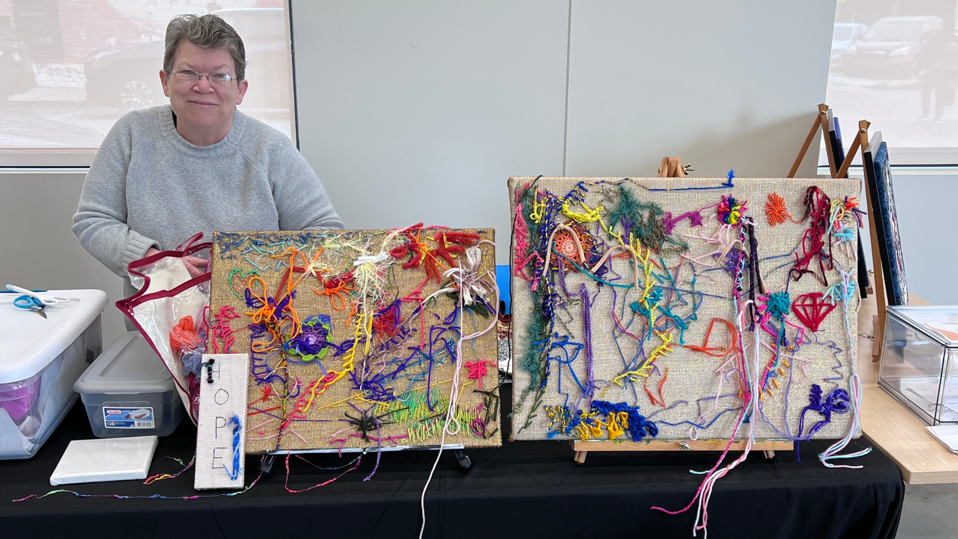 A woman smiling behind a table displaying fiber art that she has created.