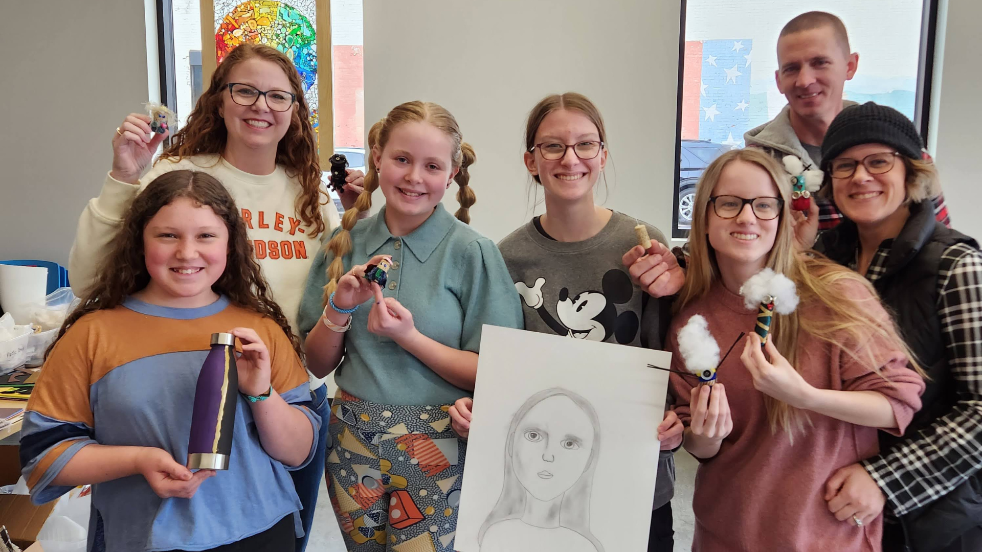 A group of middle-school aged girls with their parents smiling with various arts and crafts that they made.