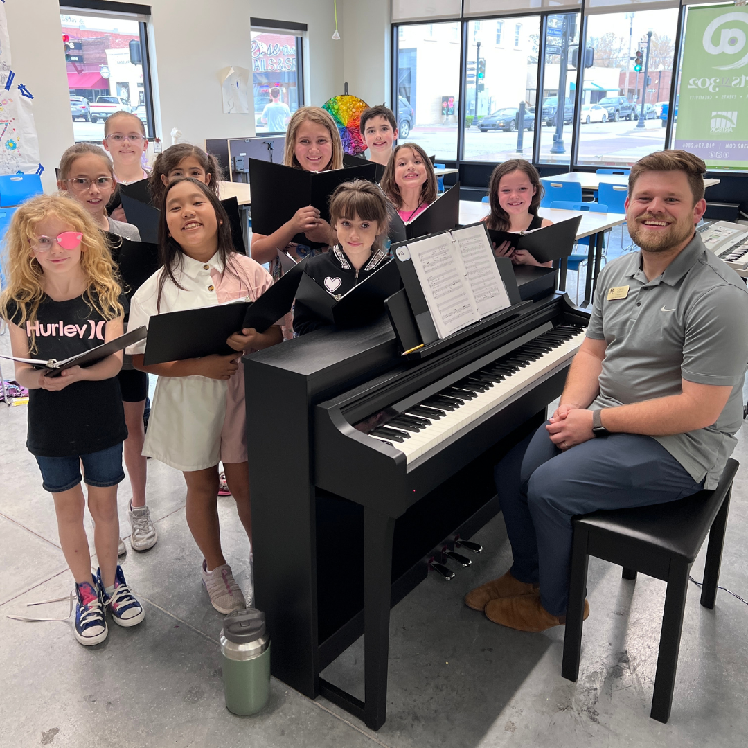 A group of children holding folders of sheet music in front of a piano and smiling. Their music instructor is smiling on the other side of the piano.